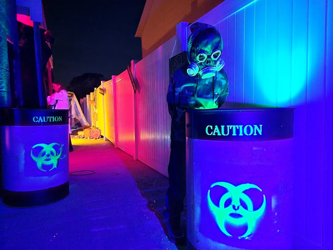 A figure in a skull gas mask leaning over a glowing CAUTION biohazard barrel, lit by blacklight in vivid blue, green and magenta.